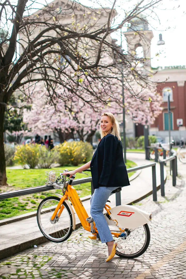 Svitlana on a yellow city bike in a Milan square, magnolias in bloom behind her.